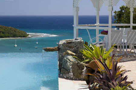 A gazebo with a view of the ocean and a swimming pool, surrounded by lush greenery and a clear blue sky.